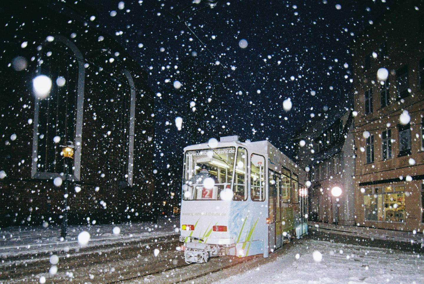 A tram is visible on a snowy street at night, with large snowflakes falling.
