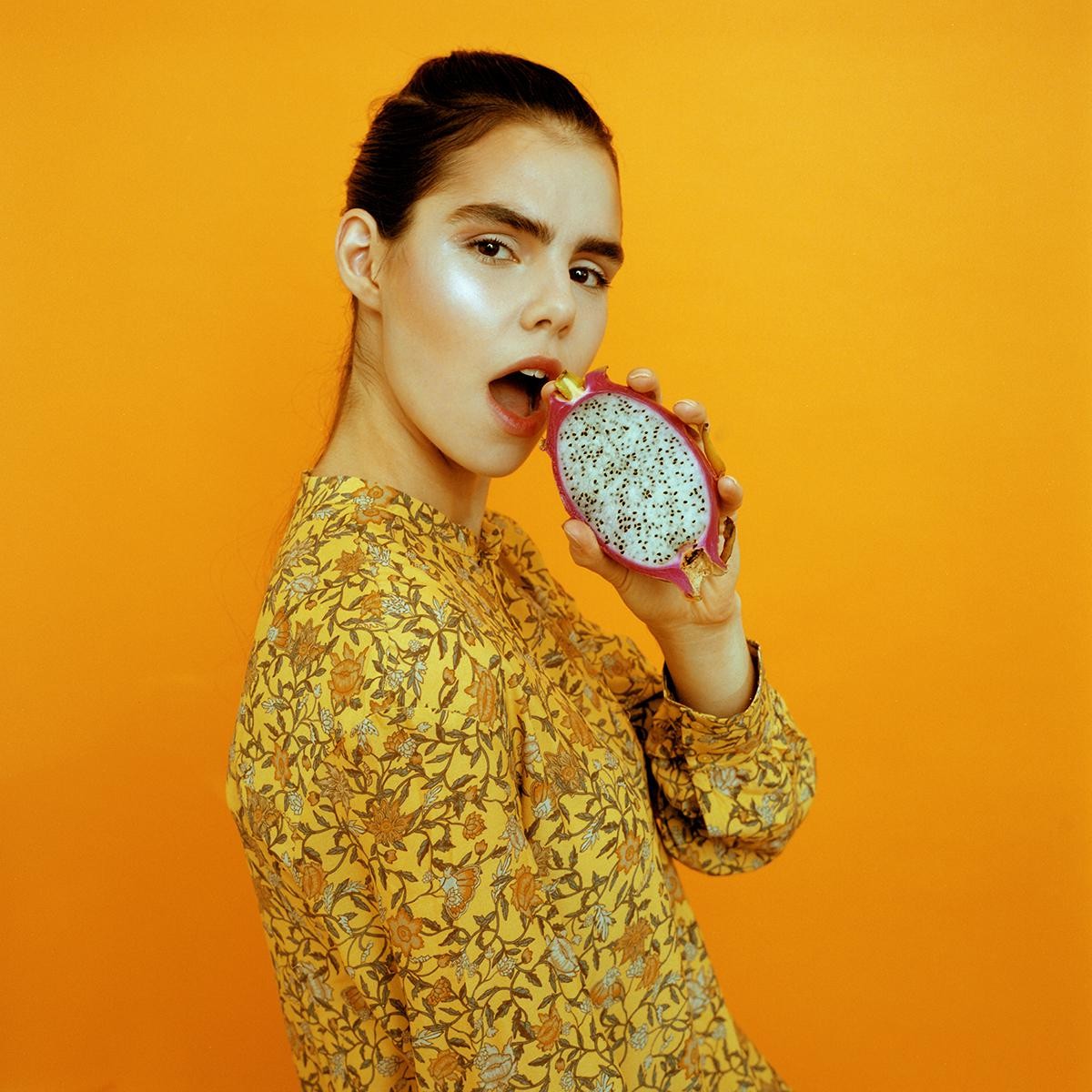 A woman in a yellow floral shirt holds a dragon fruit to her open mouth against an orange background.