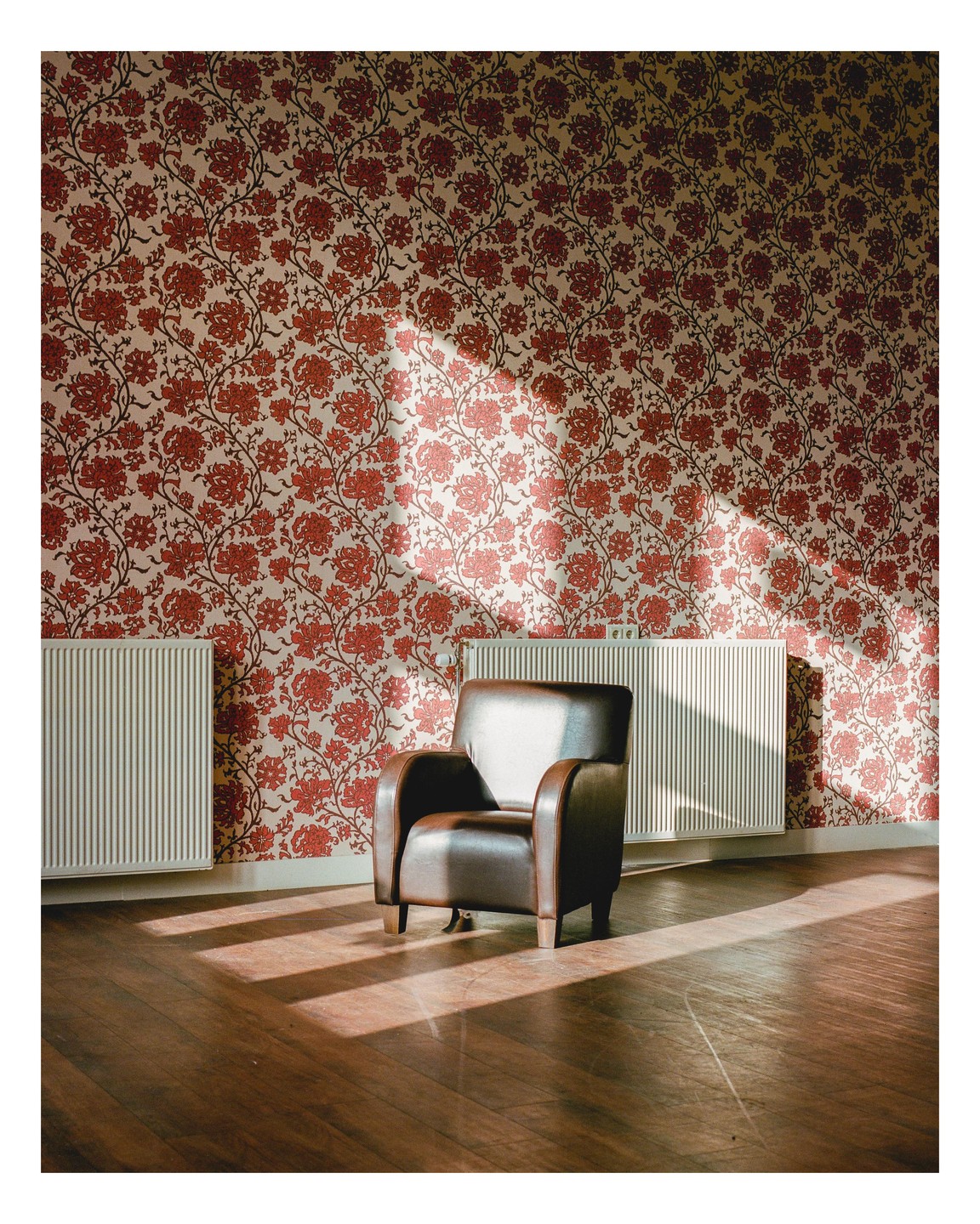 Sunlight streams into a room with floral wallpaper, illuminating a brown leather armchair and two radiators.