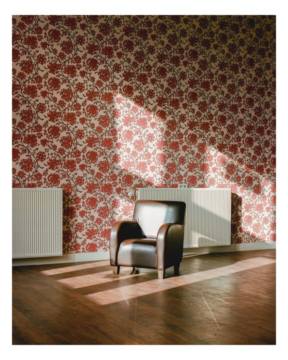 Sunlight streams into a room with floral wallpaper, illuminating a brown leather armchair and two radiators.