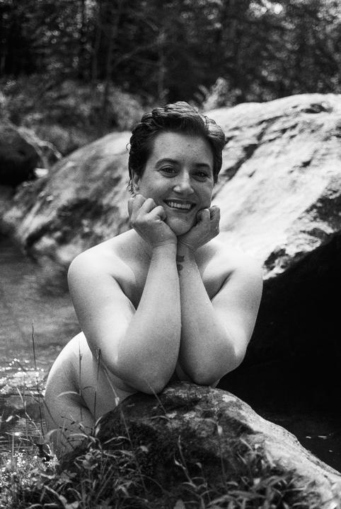 A smiling woman with short hair poses in water, resting her chin on her hands, with rocks and foliage in the background.