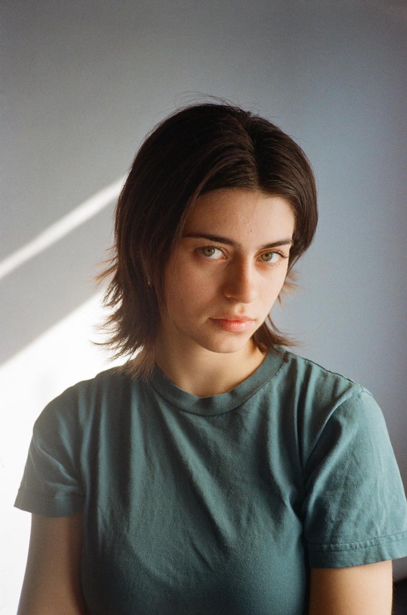A woman with short hair and a green shirt looks directly at the viewer, with a diagonal shadow on the wall behind her.