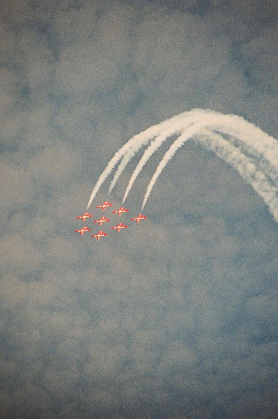 Seven red and white airplanes fly in formation, leaving white smoke trails in a cloudy sky.