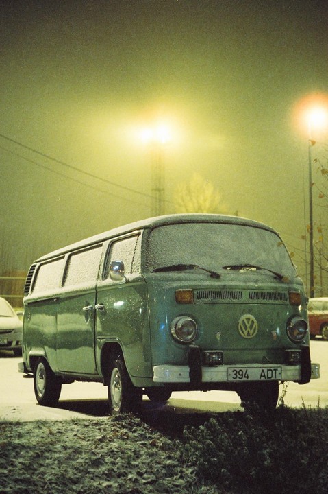 A green Volkswagen van is parked in the snow at night under a street light.