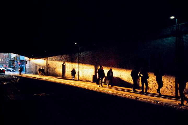 People walk along a sidewalk next to a brick wall illuminated by golden light at night.