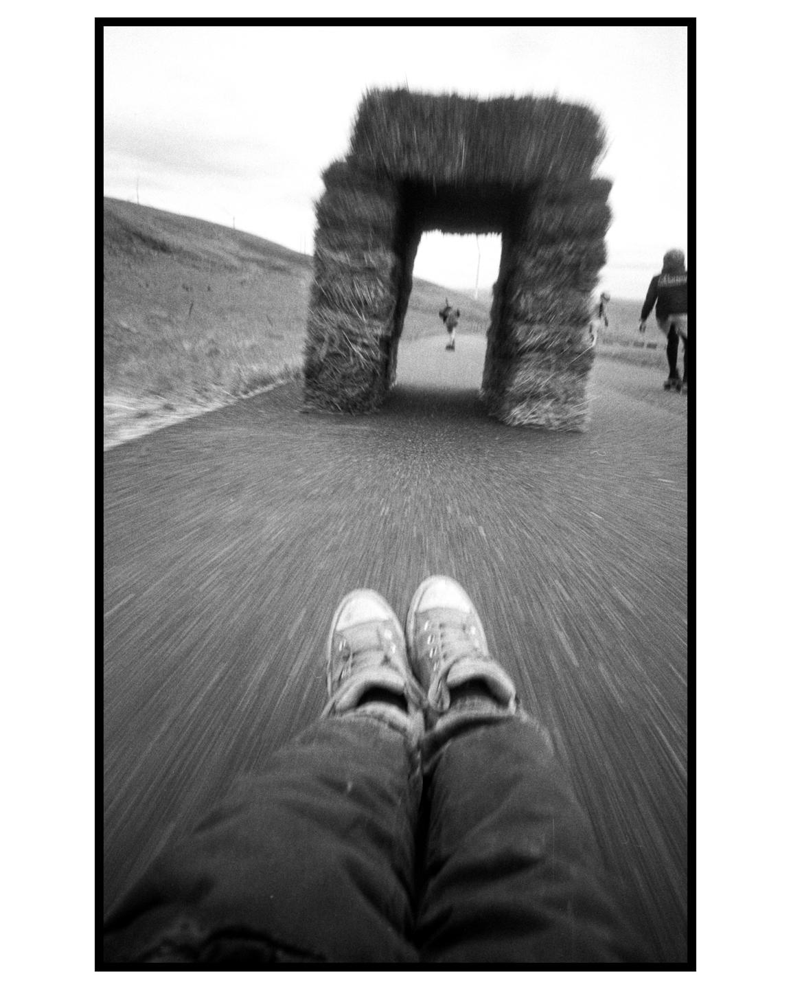 A person's legs and shoes are visible in the foreground as they ride a street luge towards a hay bale archway.