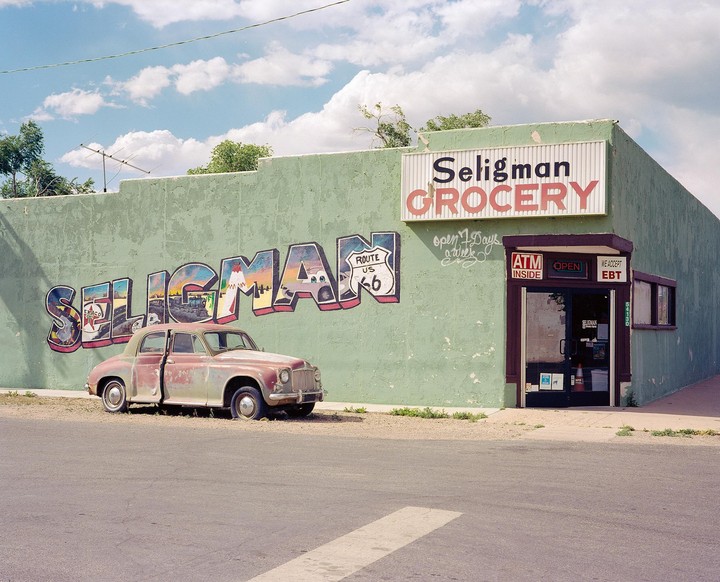 A faded green grocery store with a large mural of the word "Seligman" and a vintage car parked in front.
