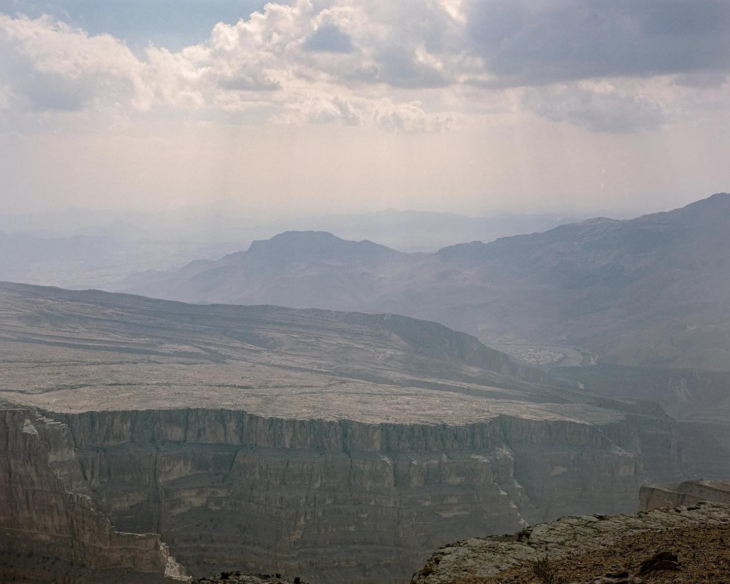 A vast canyon with a flat plateau on one side and layered mountains in the distance is visible under a cloudy sky.