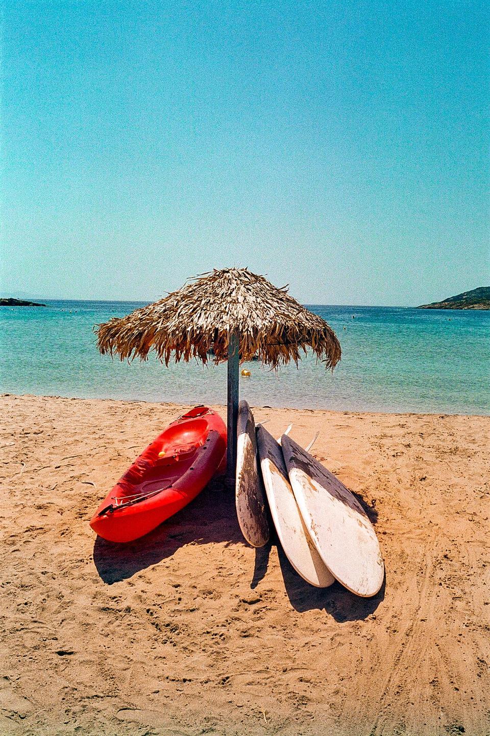A red kayak and three surfboards rest on a sandy beach under a straw umbrella, with the Aegean Sea and distant land visible in the background.