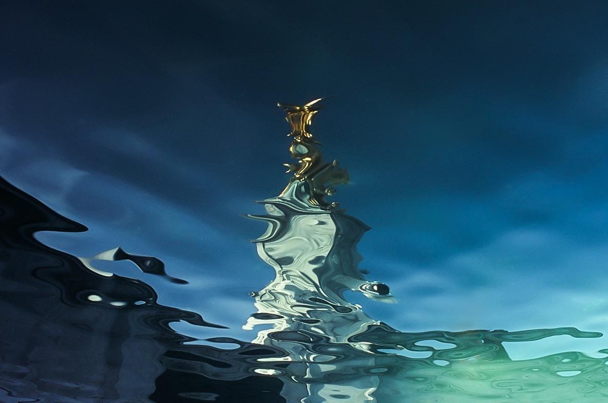 A golden statue is reflected in rippling water against a blue sky.