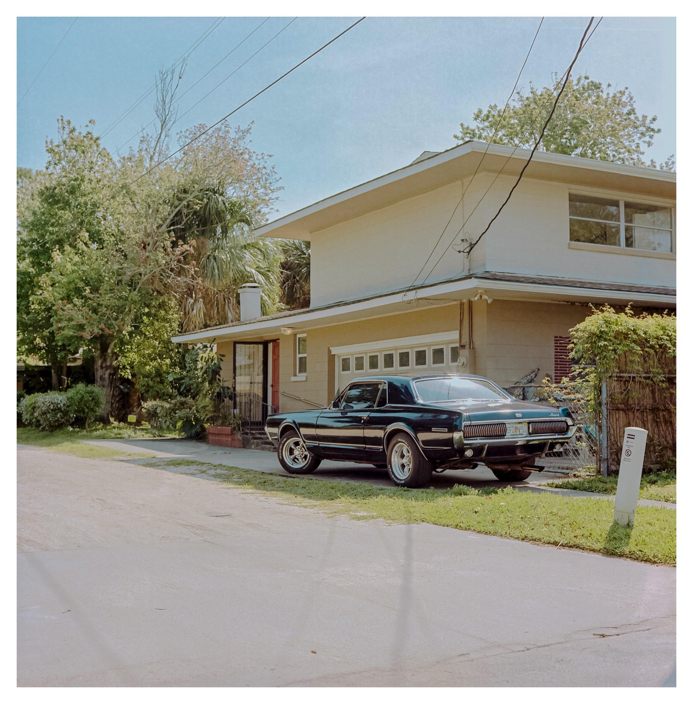 A black Mercury Cougar is parked on a grassy verge next to a house with a garage.