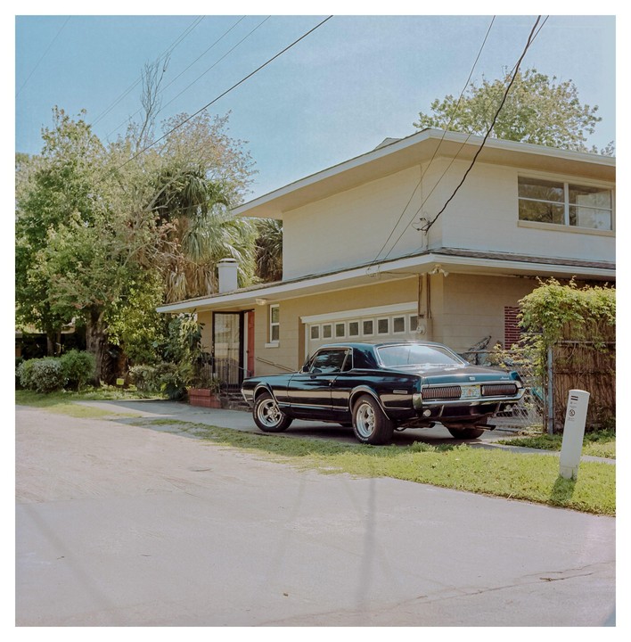 A black Mercury Cougar is parked on a grassy verge next to a house with a garage.