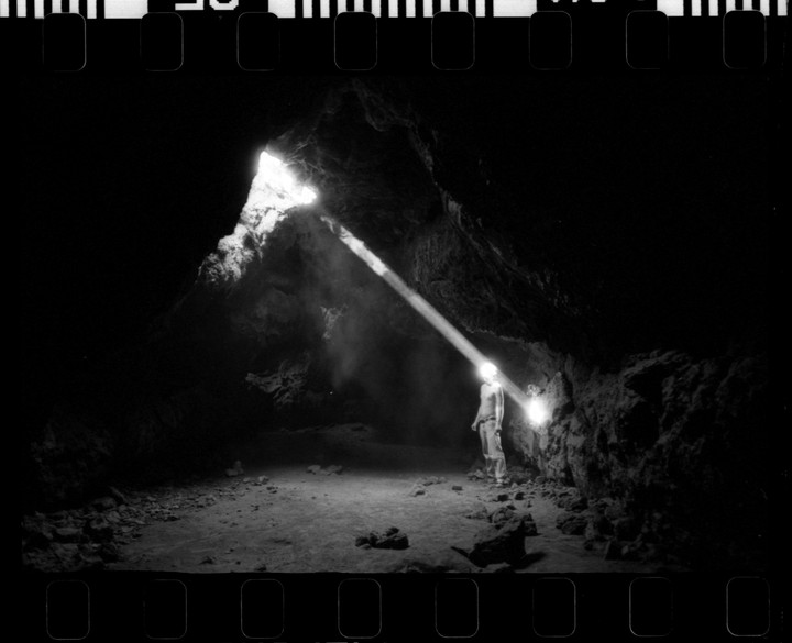 A man stands in a dark cave illuminated by a beam of light from above and a headlamp.