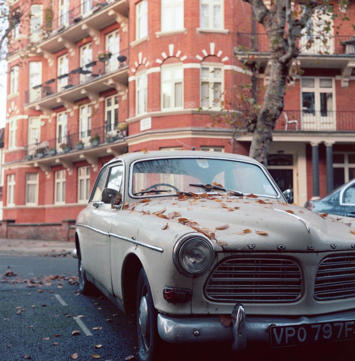 A vintage car covered in autumn leaves is parked on a street in front of a red brick building.