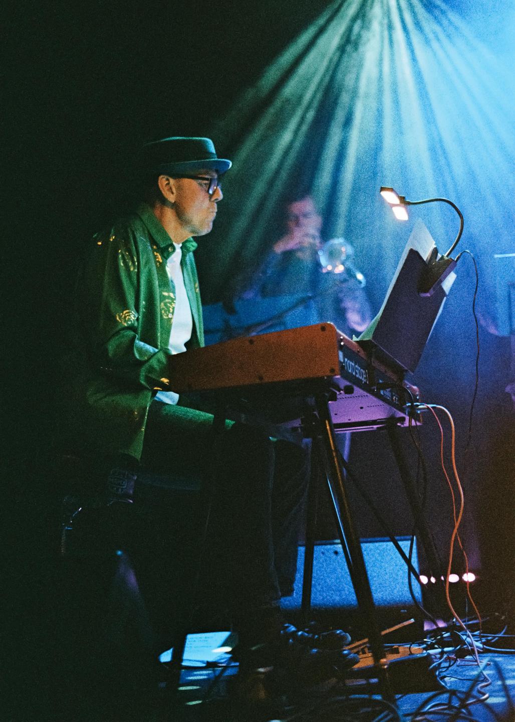 A man in a green shirt and hat plays a keyboard on a stage with blue stage lighting.