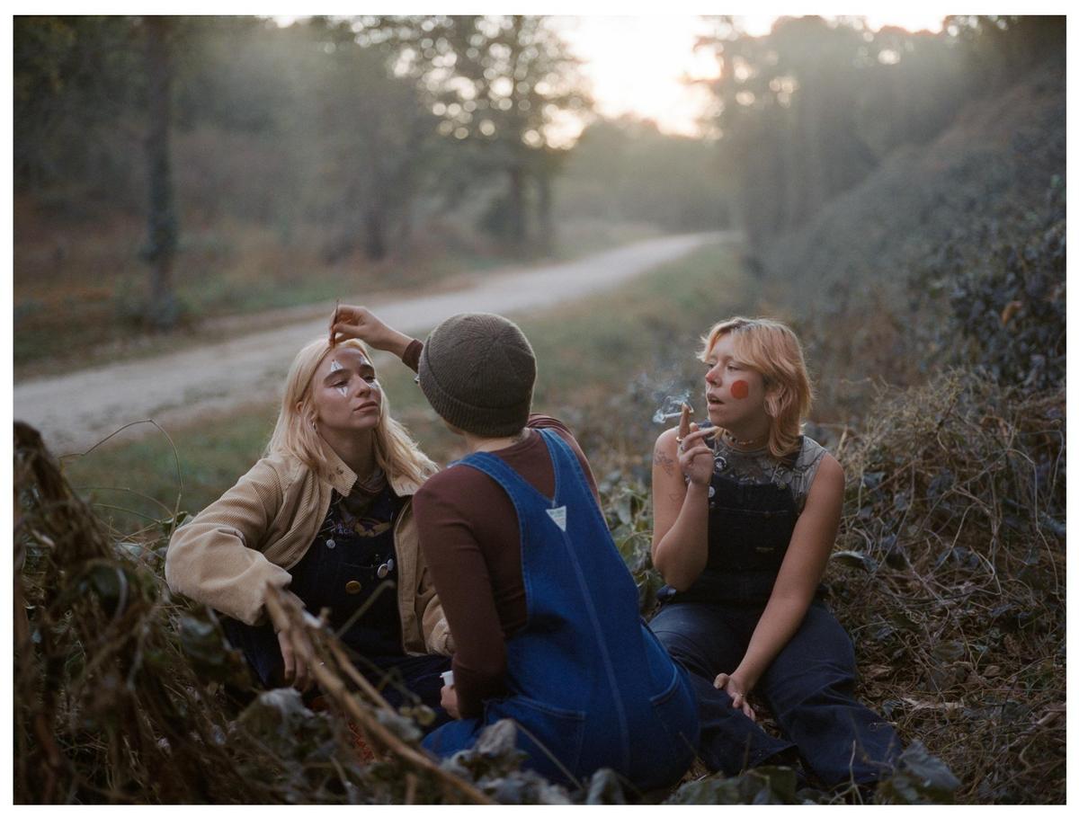 Three young people are sitting on the ground in a rural setting, with one applying makeup to another while the third smokes a cigarette.