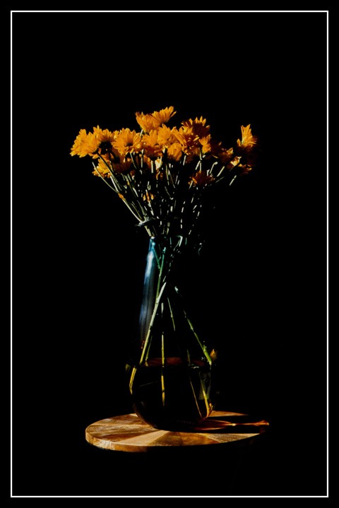 A bouquet of orange flowers in a glass vase sits on a wooden surface against a dark background.