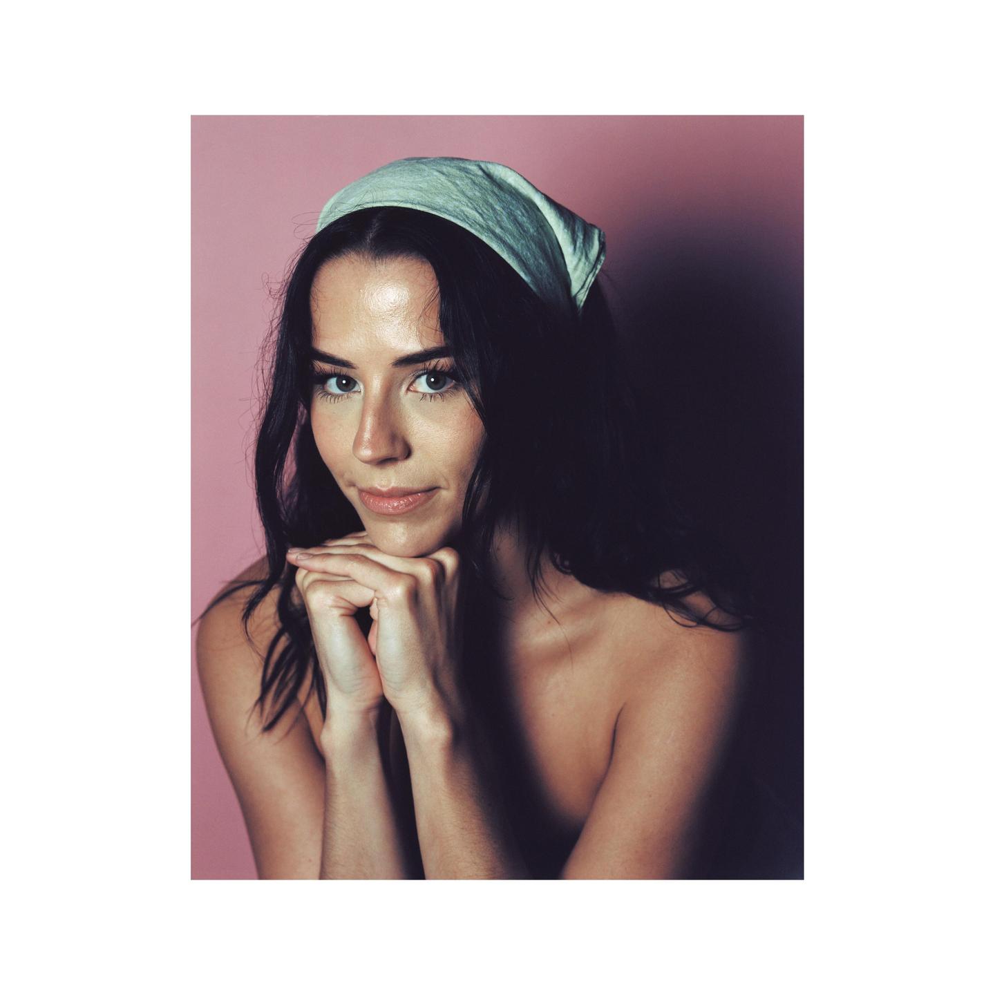 A woman with dark hair and a light-colored headband rests her chin on her clasped hands, looking directly at the viewer against a pink background.