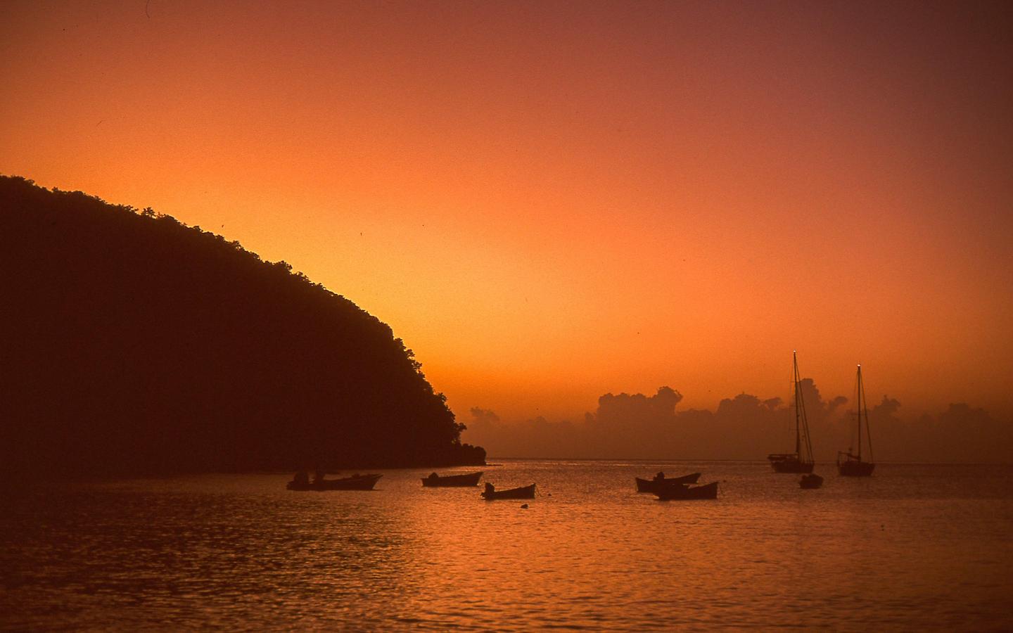 A body of water with several boats is silhouetted against a vibrant orange and red sky with a dark hillside on the left.