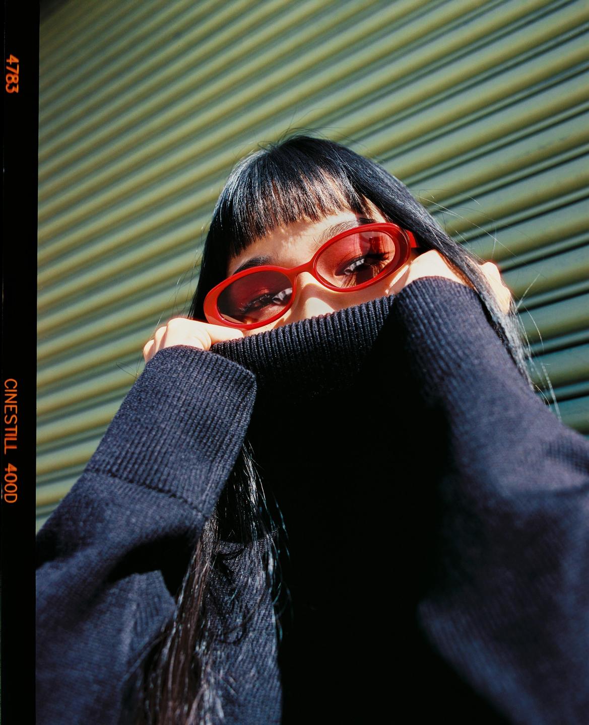 A woman wearing red sunglasses and a dark sweater covers her face with her arms.