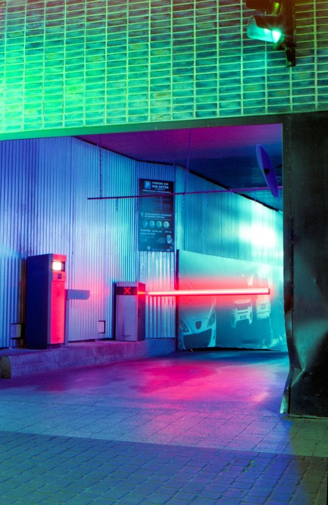 A parking garage entrance is illuminated by neon lights, with a red barrier across the opening and a car visible inside.