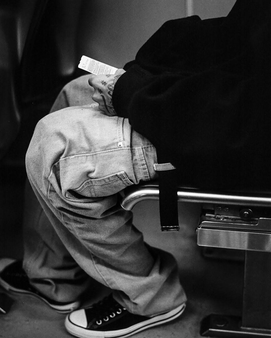 A person wearing jeans and sneakers sits on a seat, holding a small piece of paper in a tattooed hand.