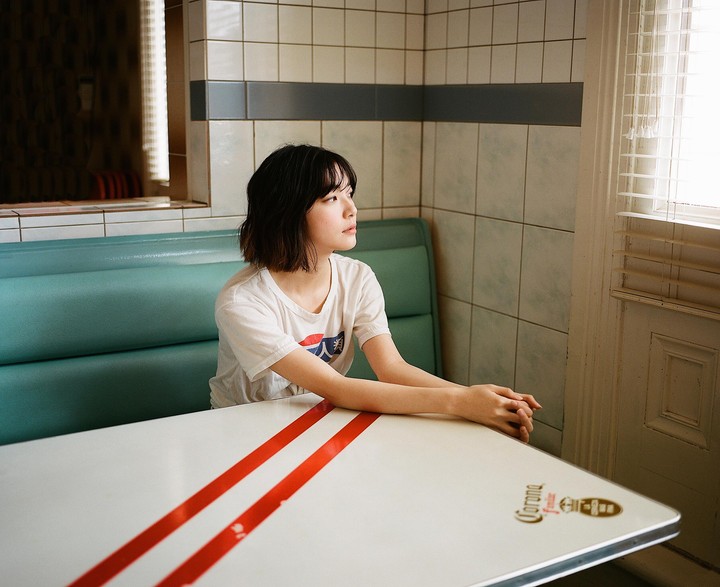 A woman sits at a diner booth with a white table that has two red stripes and a Corona logo, looking out a window with blinds.