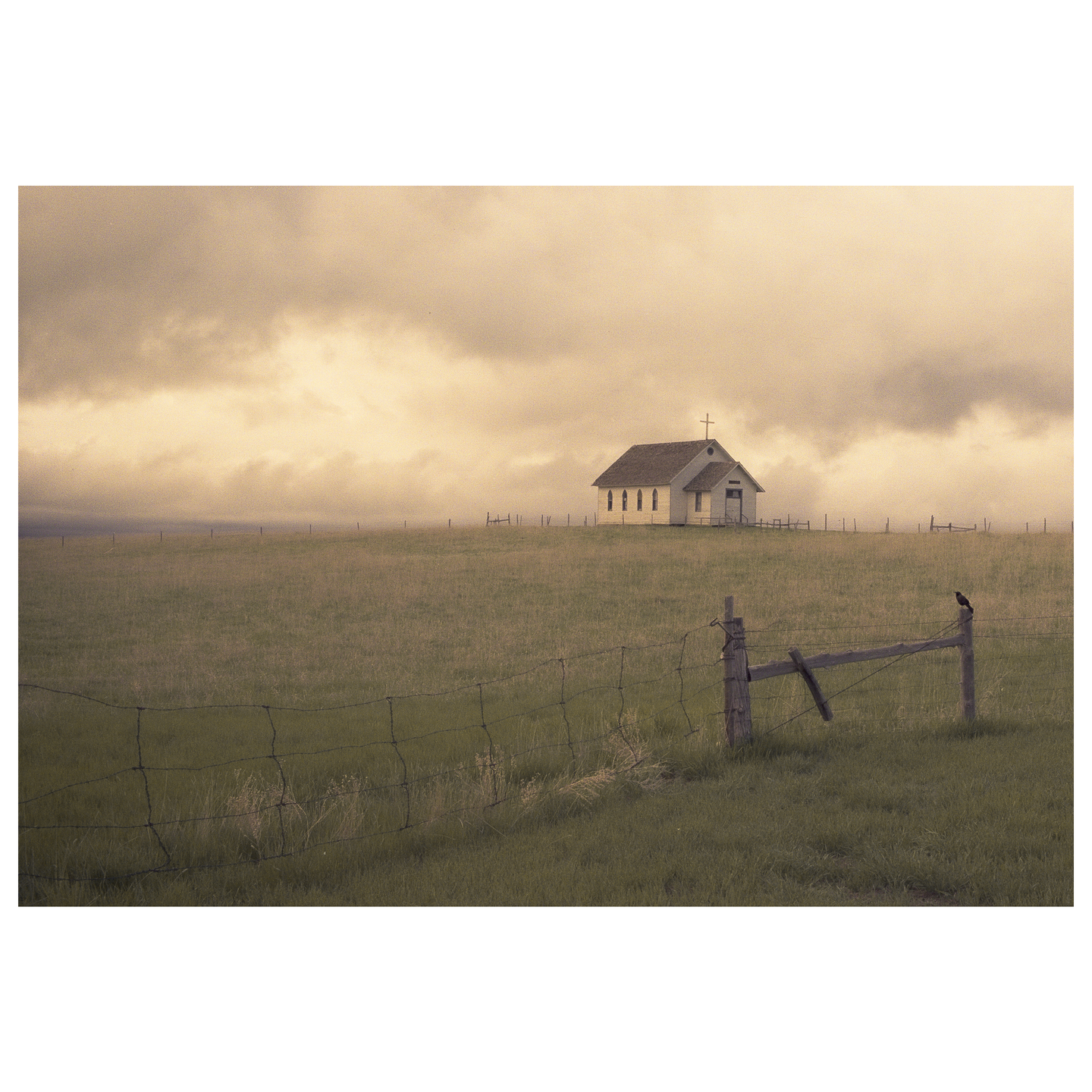 A small white church with a cross on its roof stands in a grassy field under a cloudy sky, with a bird perched on a fence post in the foreground.