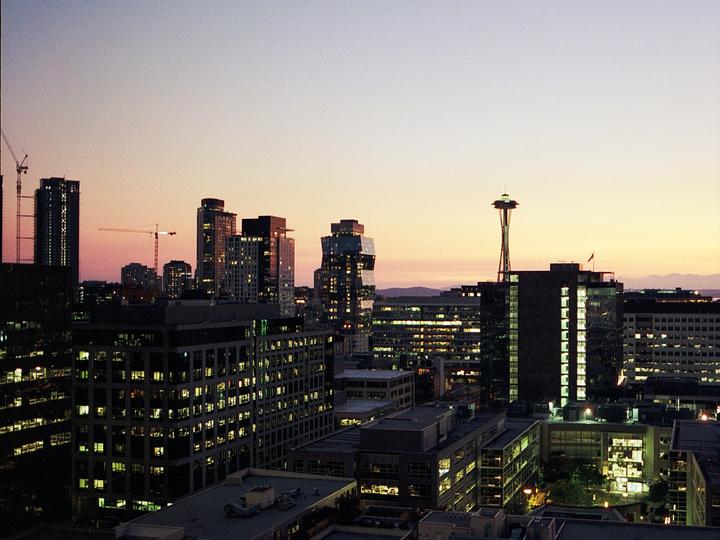 A cityscape of Seattle at dusk shows the Space Needle and other buildings with illuminated windows against a colorful sky.