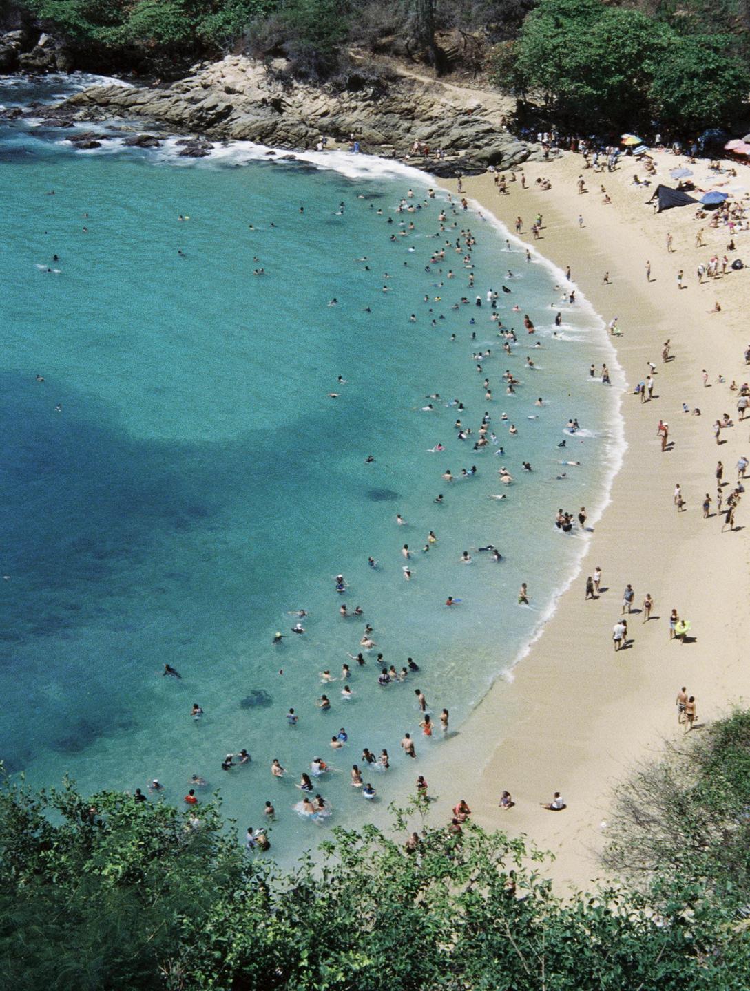A crowded beach with many people swimming in the clear blue water and relaxing on the sand.