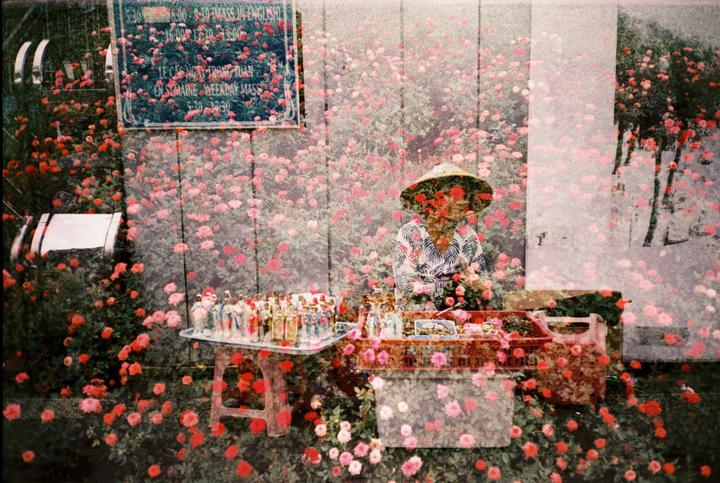 A person wearing a conical hat sits behind a stall with religious figurines and other items, all overlaid with an image of many small flowers.