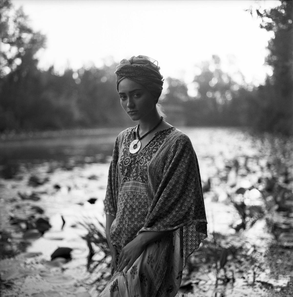 A woman wearing a patterned kaftan and a head covering stands by a body of water.