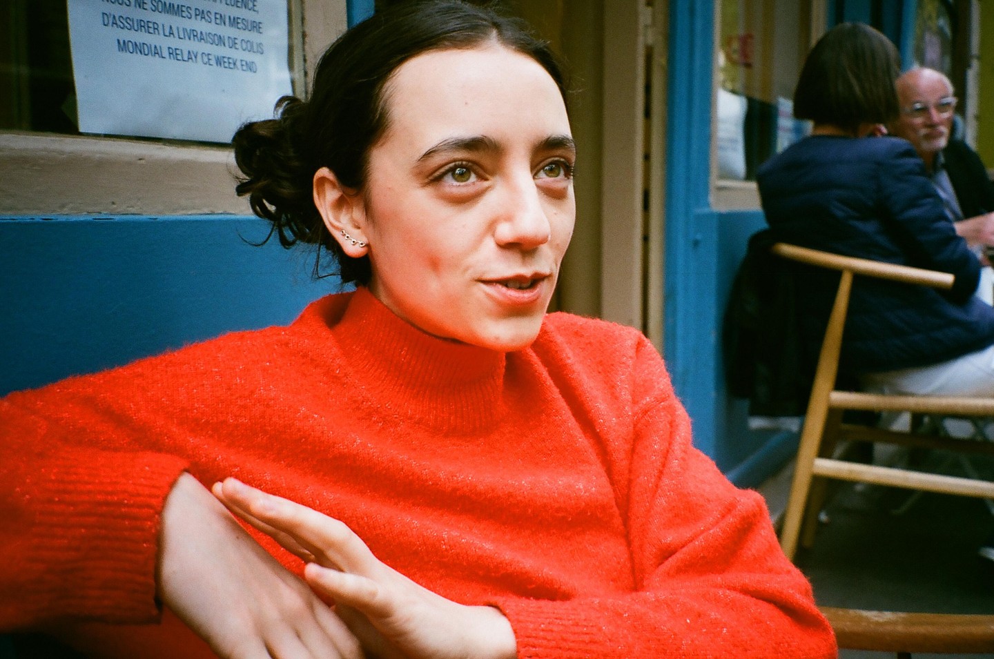 A woman in a red sweater looks to the side while sitting outdoors, with another person visible in the background.