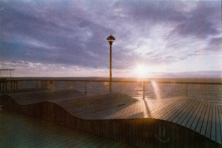 The sun sets over the ocean, casting a warm glow on a wooden boardwalk with a single lamppost.