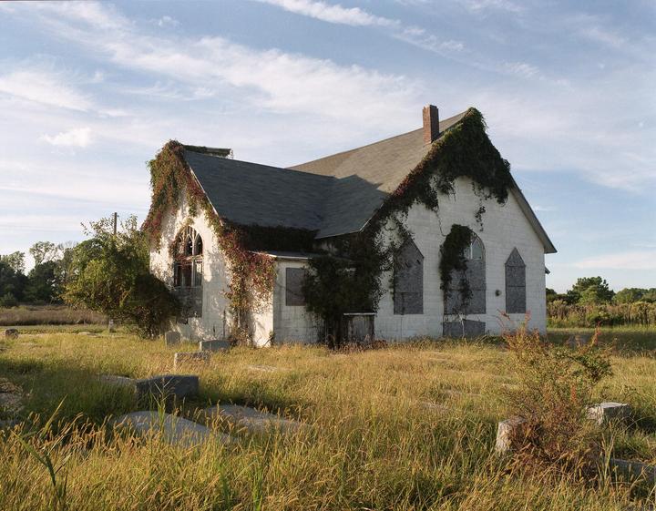An abandoned church covered in ivy stands in a field of tall grass under a partly cloudy sky.