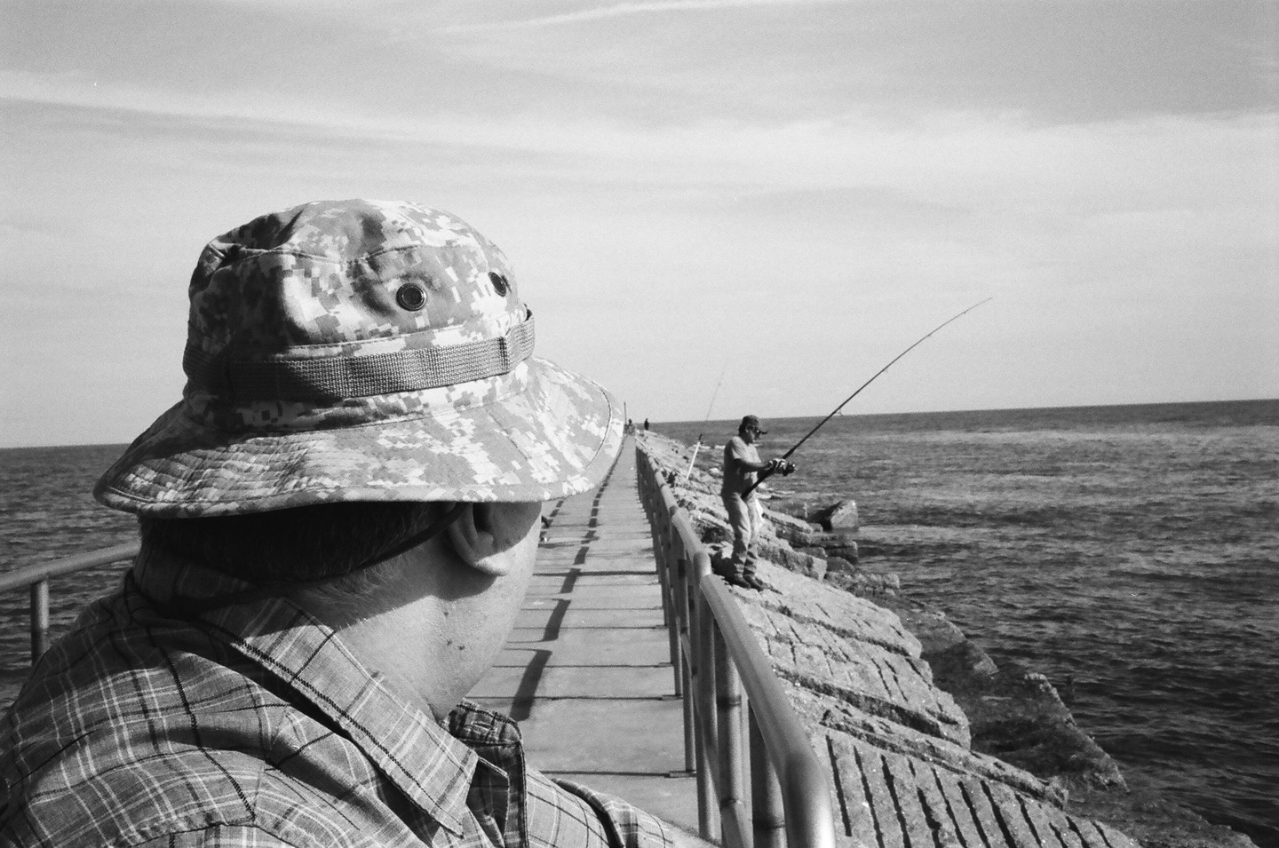 A man in a camouflage hat and plaid shirt looks out at the ocean where another man is fishing from a rocky pier.