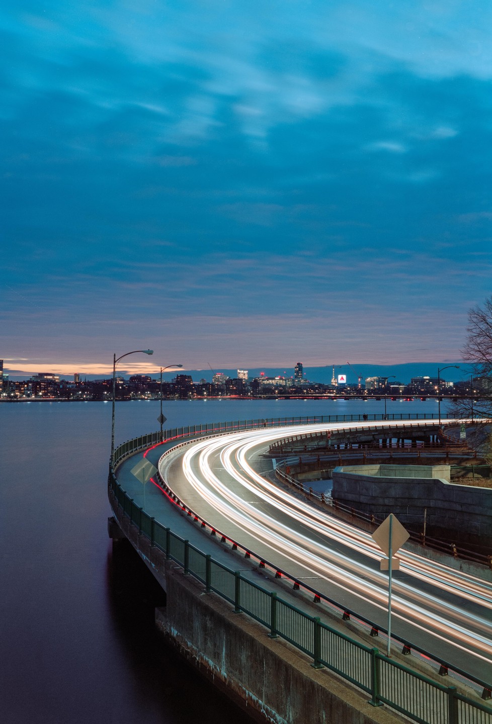 A long exposure captures light trails from vehicles on a curved highway next to a body of water with a city skyline in the distance at dusk.