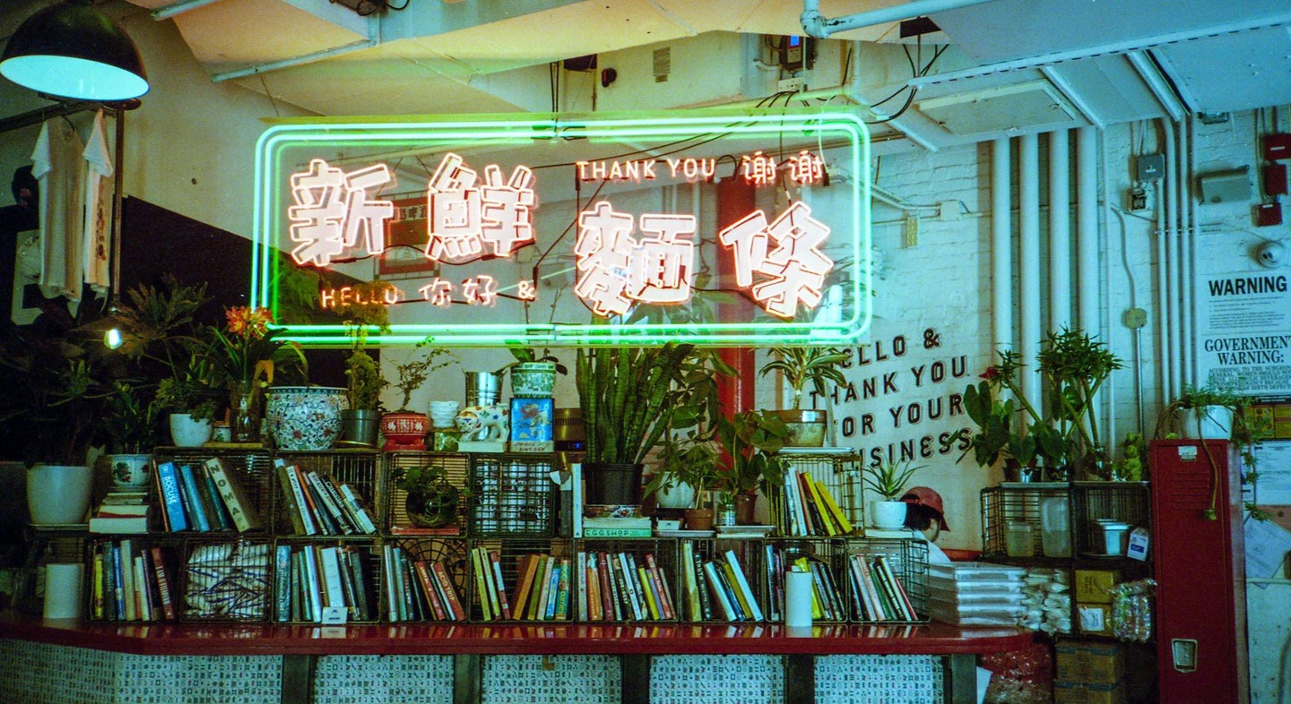 A neon sign with Chinese characters and English text hangs above a counter filled with books and plants.