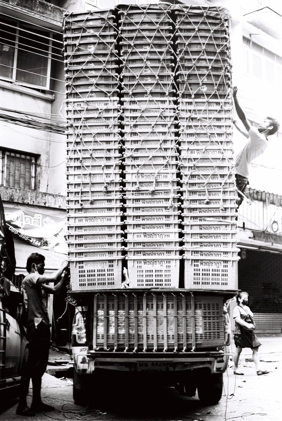 Two men are securing a very tall stack of crates on the back of a truck, while a woman walks by.