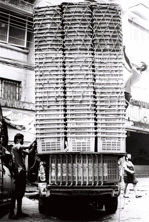 Two men are securing a very tall stack of crates on the back of a truck, while a woman walks by.