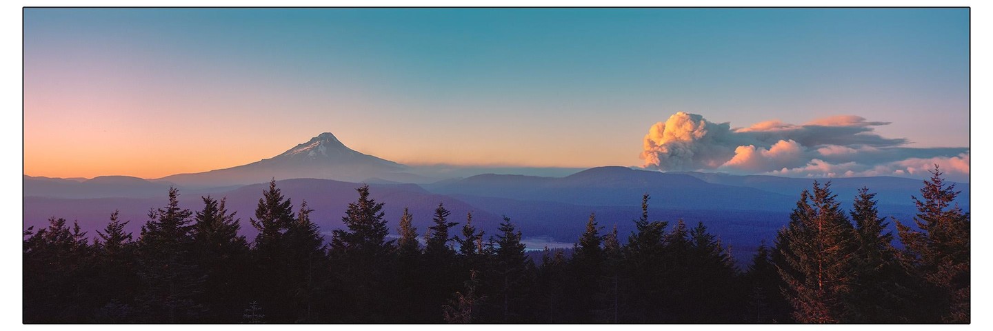 A panoramic landscape shows a mountain peak and a large cloud of smoke rising from behind a forest of evergreen trees at sunset.