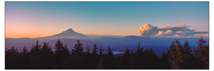 A panoramic landscape shows a mountain peak and a large cloud of smoke rising from behind a forest of evergreen trees at sunset.