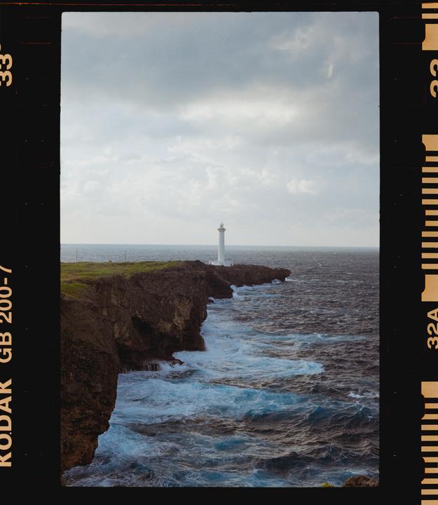 A white lighthouse stands on a rocky cliff overlooking a turbulent ocean under a cloudy sky.