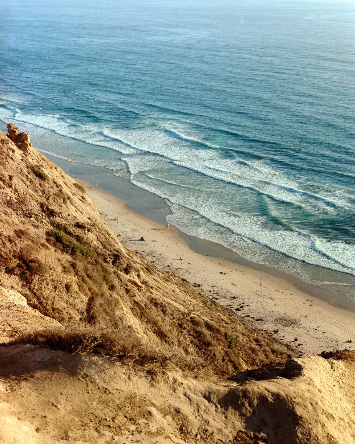 A steep, sandy cliff overlooks a beach with gentle waves rolling onto the shore.