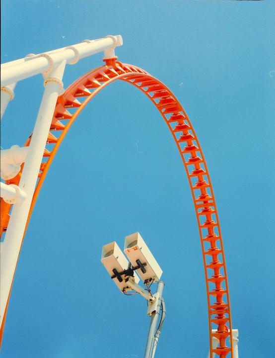 An orange and white roller coaster track curves against a clear blue sky, with two white security cameras mounted on a pole in the foreground.