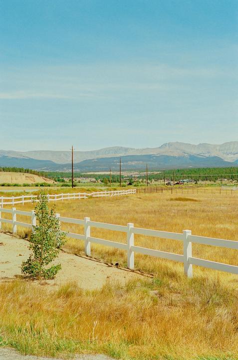 A white fence runs through a field of dry grass with mountains in the distance under a clear sky.
