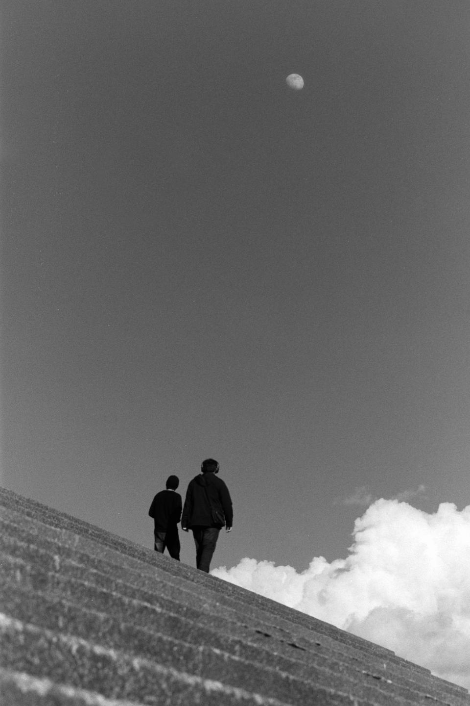 Two men walk up a steep, textured incline under a vast sky with a visible moon and a cloud.