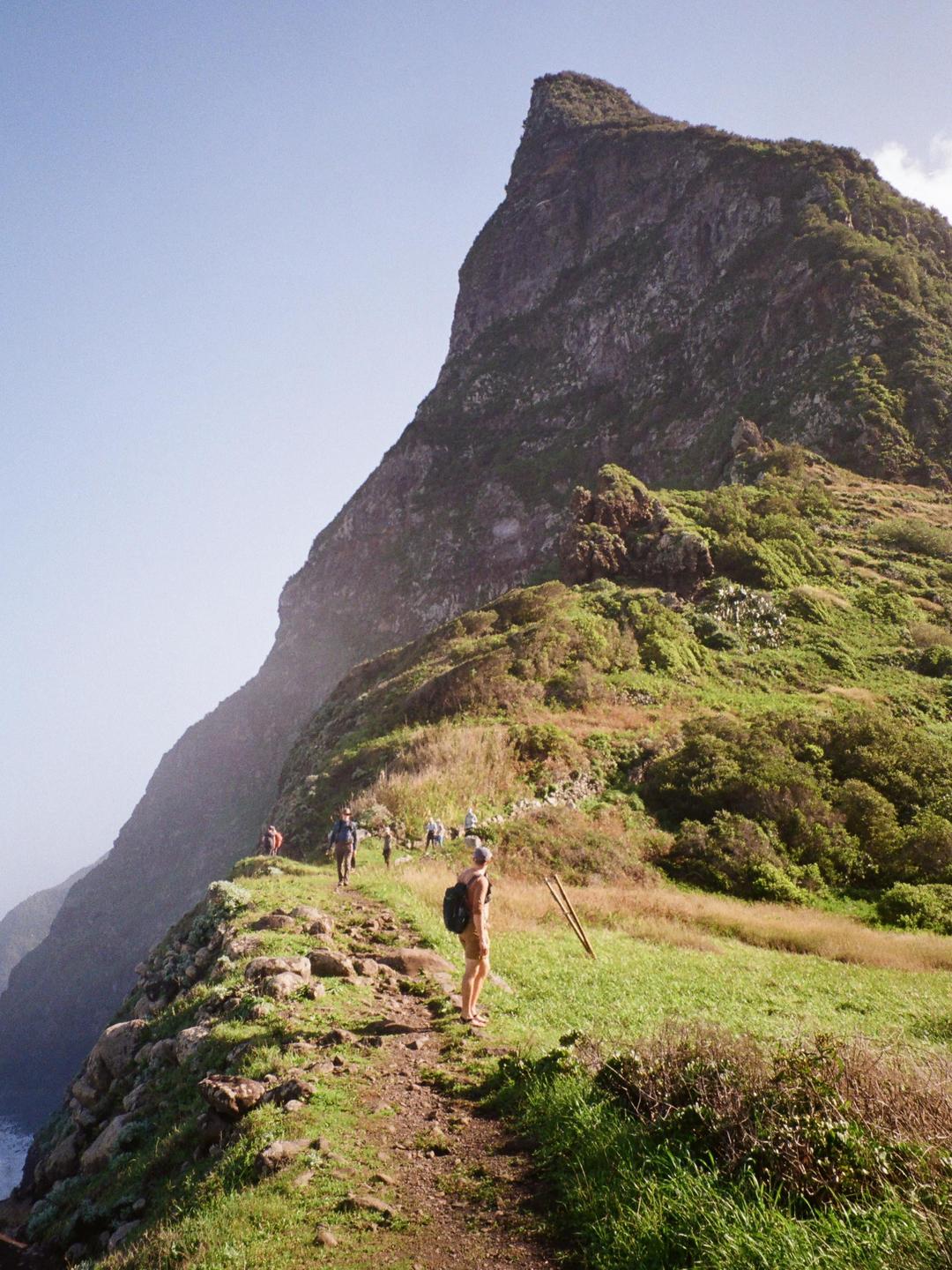 A group of hikers walks along a narrow path on a steep, green mountainside overlooking the ocean.