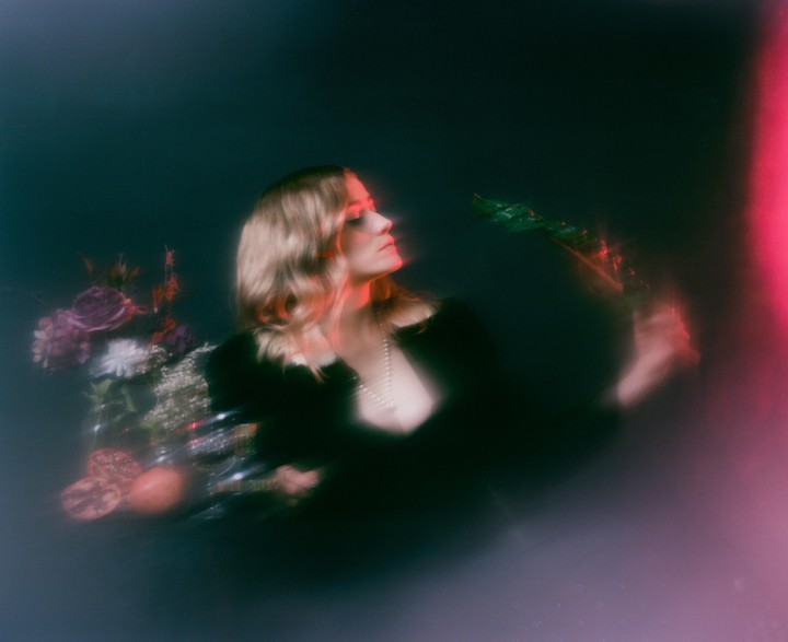 A woman with blonde hair and a pearl necklace holds a green branch while sitting next to a bouquet of flowers and pomegranates.