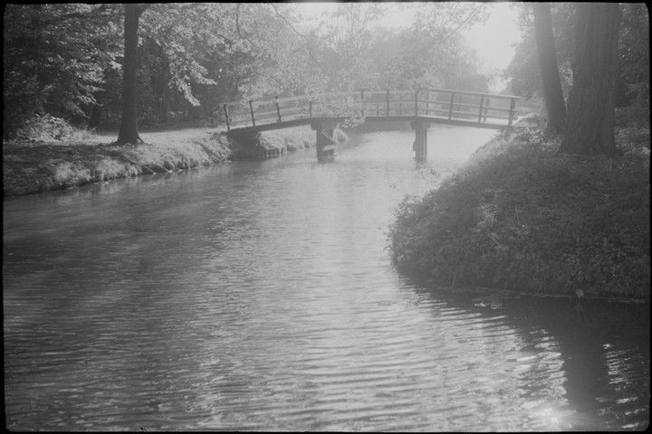 A small wooden bridge crosses a narrow river surrounded by trees and foliage.
